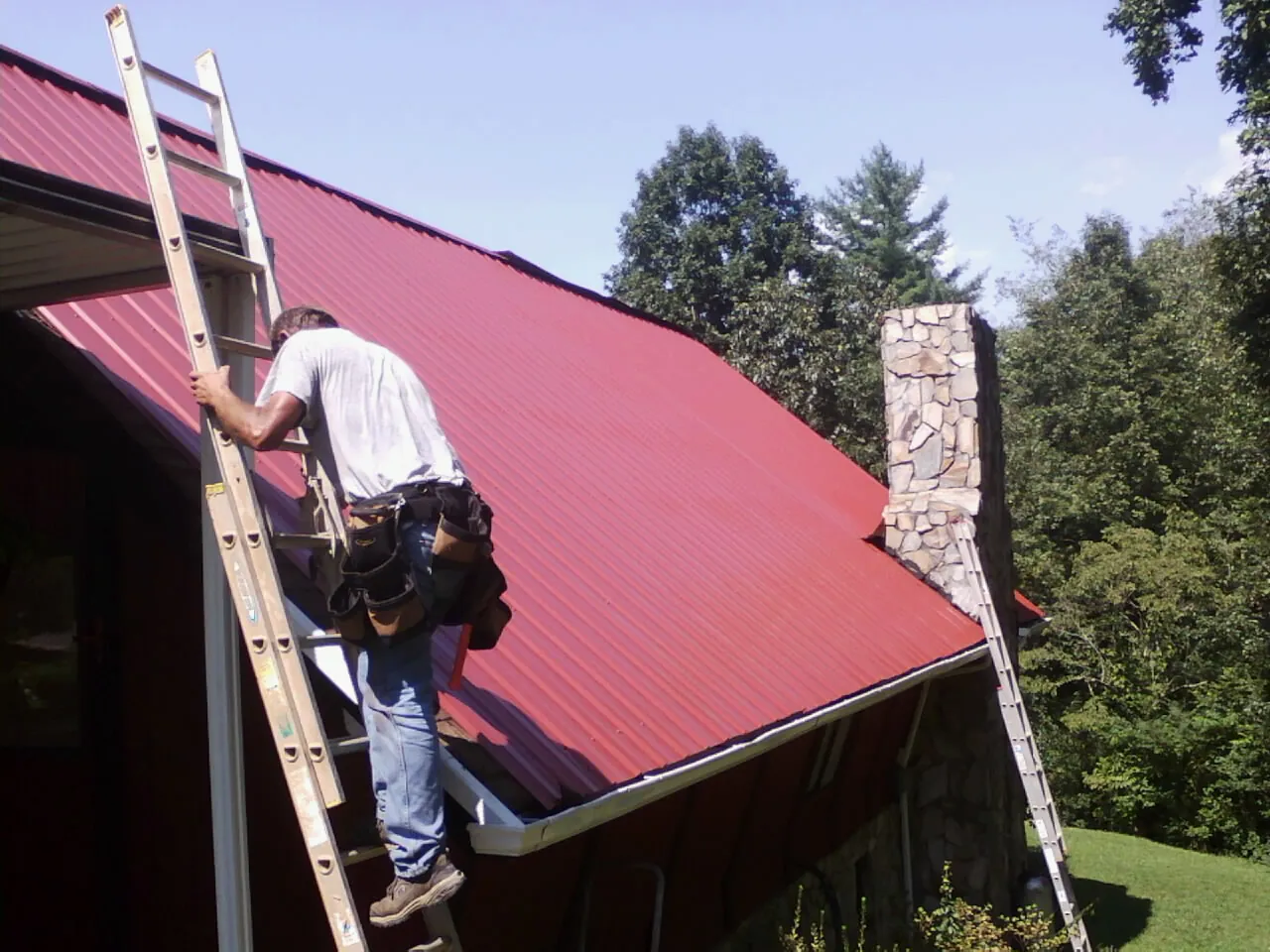 Roofer on a ladder beside a red metal roof and stone chimney — Soffit & Fascia Repair by Roofing Brothers in Hartland